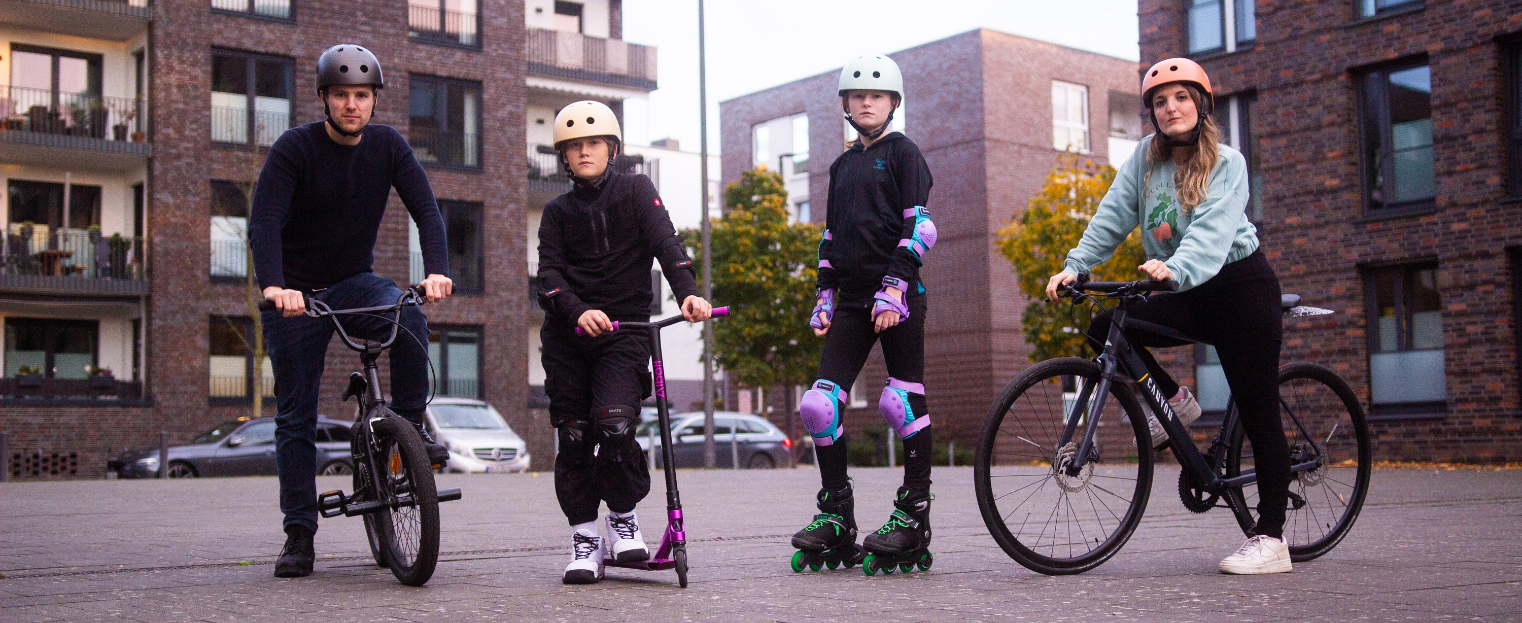 Eine Gruppe aus Action Sport-Athleten in Schutzausrüstung bestehend aus  Helm, Knieschonern und Ellbogenschonern stehen auf einem Platz in einer Stadt.