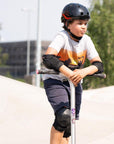 Ein Junge mit einem Scooter steht im Skatepark ausgerüstet mit Skateschutzausrüstung